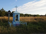 Na zdjęciu przydrożna kapliczka w kolorze niebieskim na tle złotych łanów zbóż