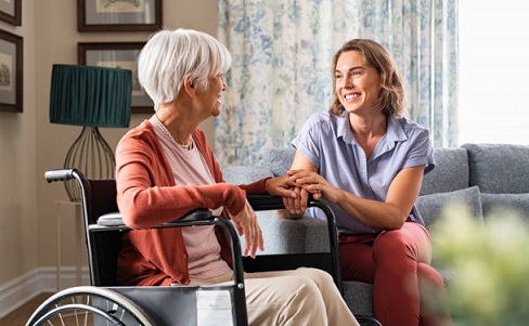 zdjęcie przedstawia starszą panią na wózku, przy niej siedzi młoda kobieta, która się uśmiecha i trzyma starszą panią za rękę