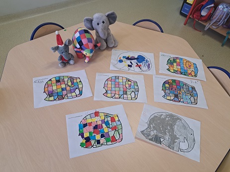 Zdjęcie przedstawia prace plastyczne na stoliku szkolnym. Prace przedstawiają słonie. Na stoliku znajdują się również pluszaki słonie.